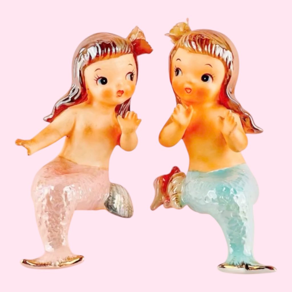 A pair of vintage mid-century Norcrest mermaid figurines shelf sitters with  blue and pink tail, soft gold accents against a soft pink background.
