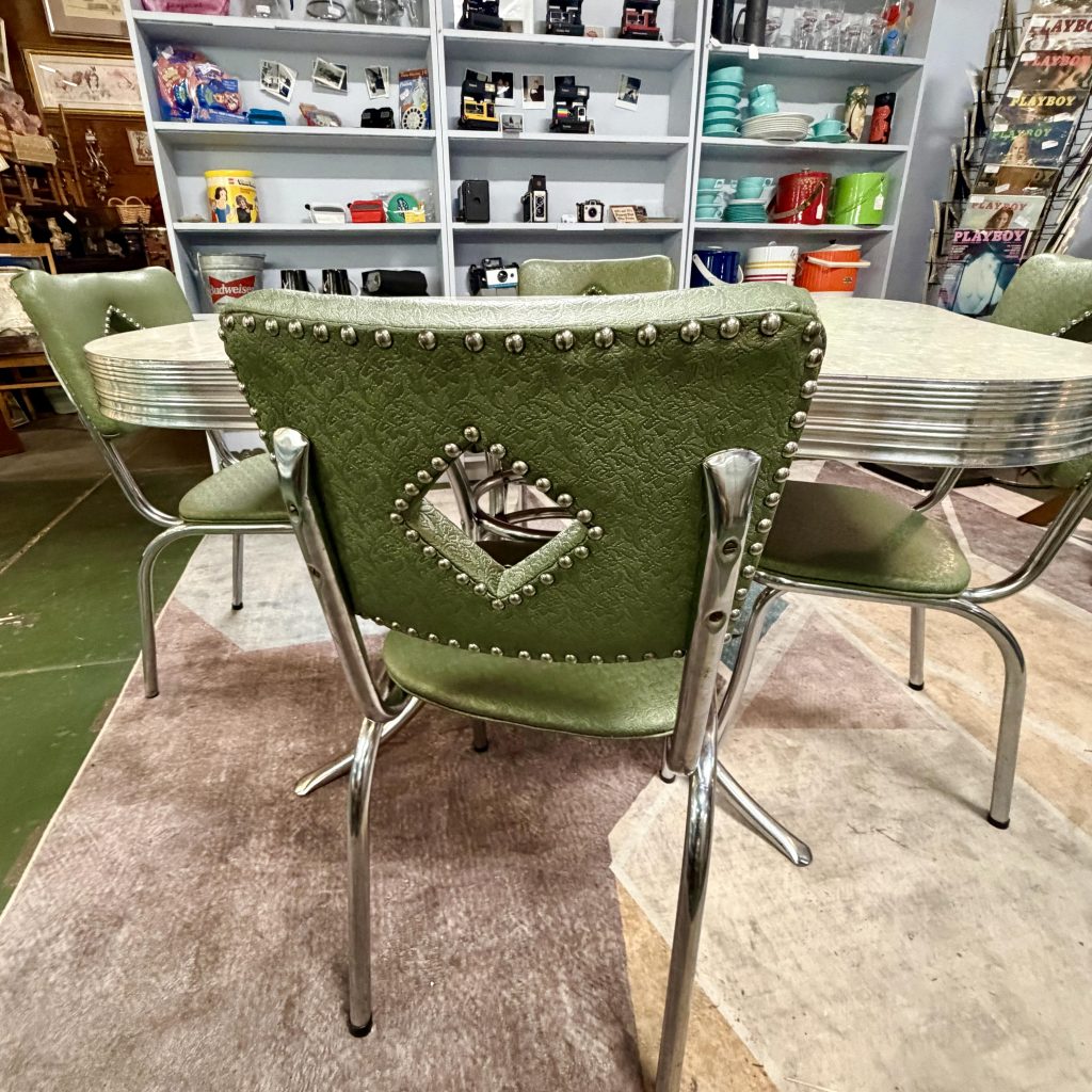 Close-up of a vintage mid-century modern dining chair featuring avocado green textured vinyl upholstery, a diamond-shaped cutout back with chrome nailhead trim, and a tubular chrome frame.
